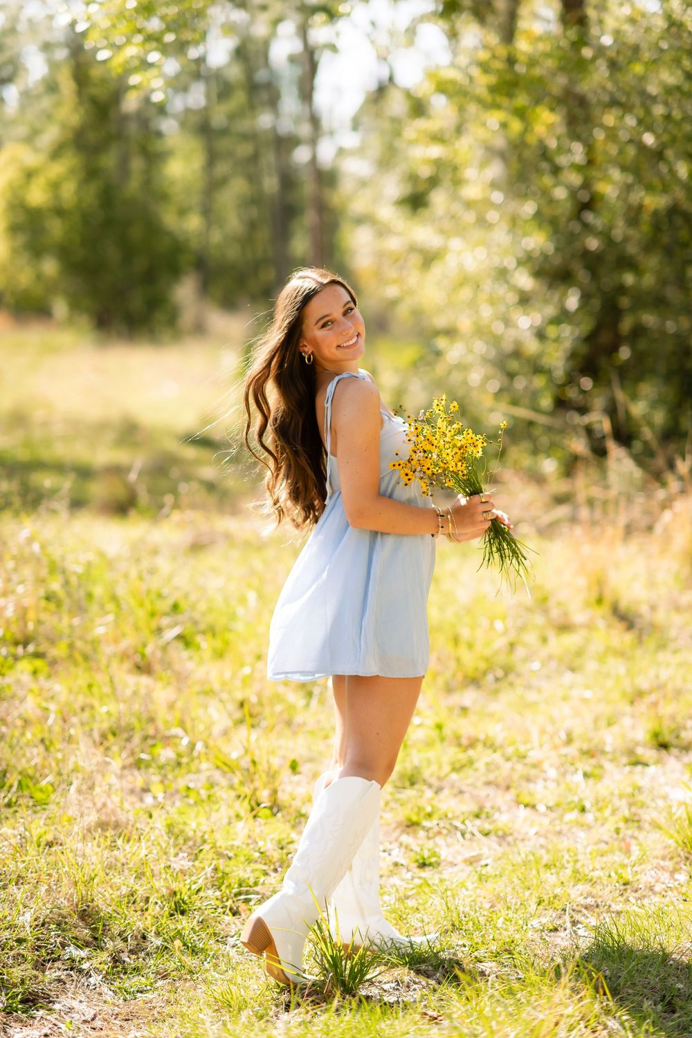 Young woman in blue dress and white boots holding yellow wildflowers, smiling outdoors in sunny green meadow.