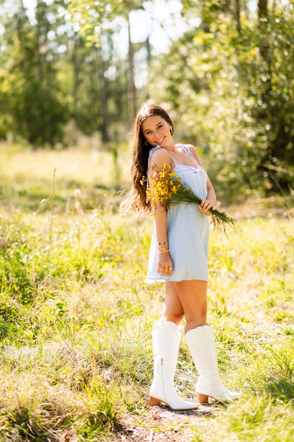 Young woman in a light blue sundress and white cowboy boots holding yellow wildflowers in a bright sunny meadow.