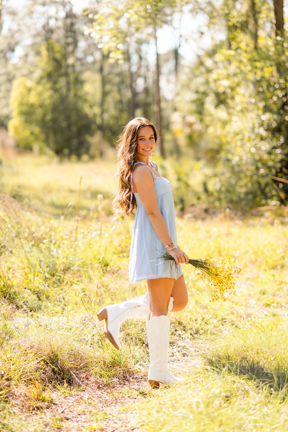Young woman in light blue dress and white cowboy boots holding yellow wildflowers in a sunny meadow.
