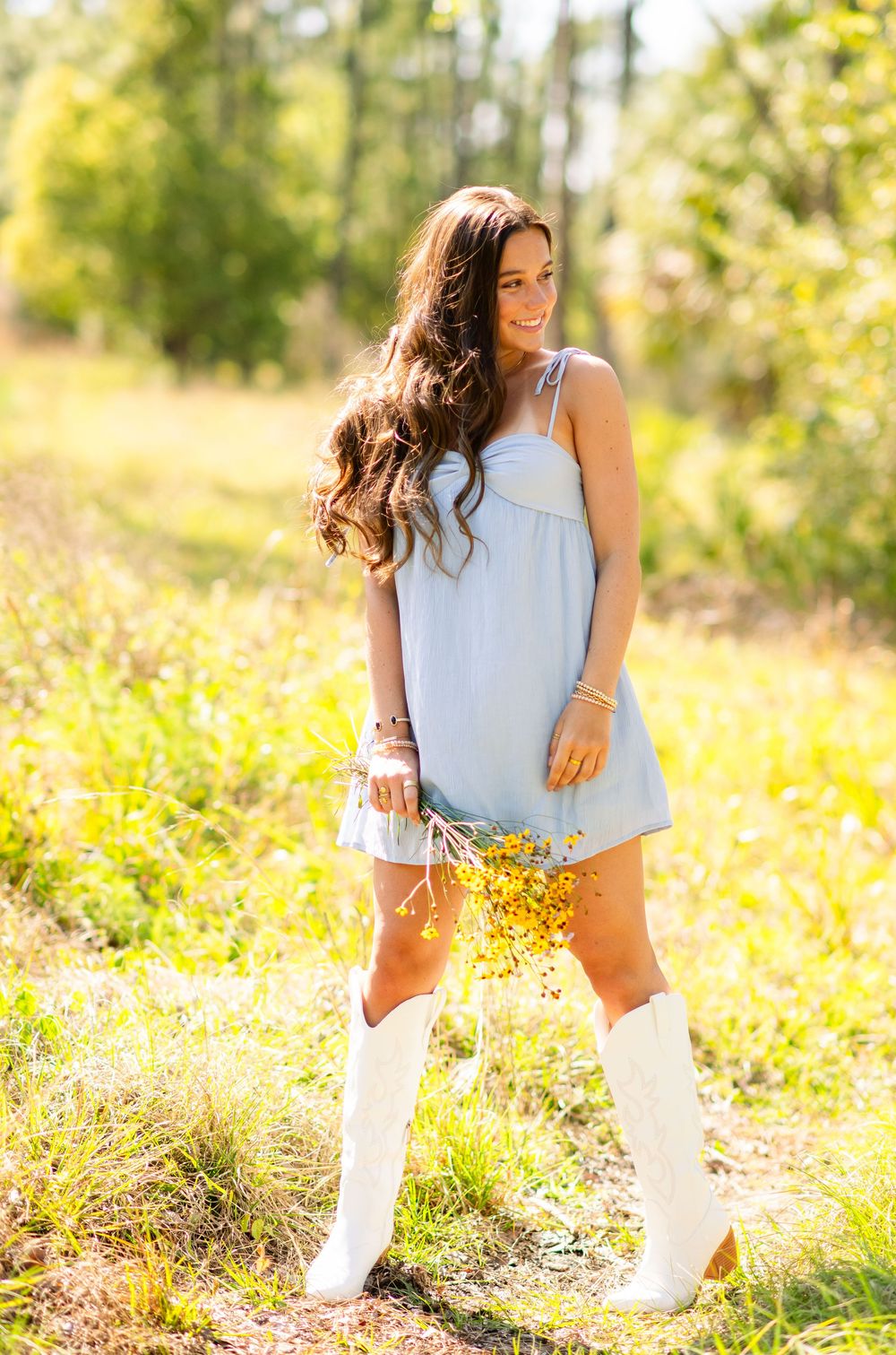 Smiling brunette in a light blue sundress and white knee-high boots holding yellow wildflowers outdoors.