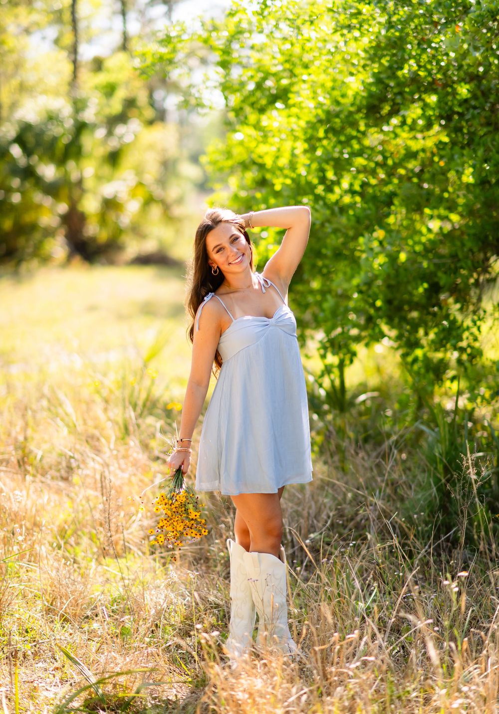 Woman in white dress holding flowers standing in sunny field with green trees in background.