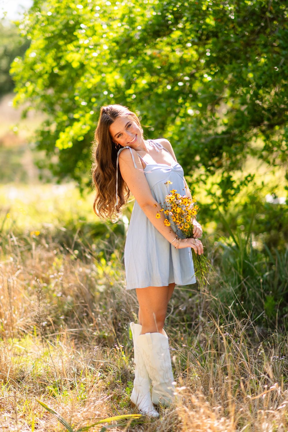 Smiling young woman in a light blue dress and white boots holding yellow wildflowers in a sunlit green field.