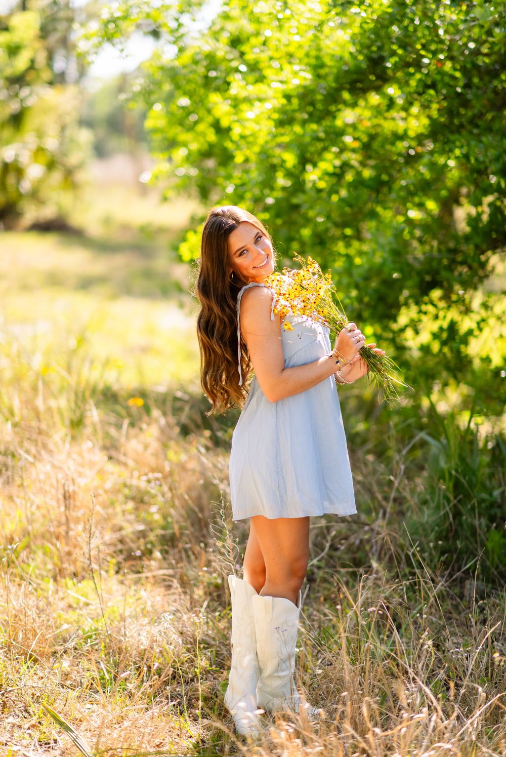 Young woman in light blue dress and white cowboy boots holding wildflowers in a sunny field.