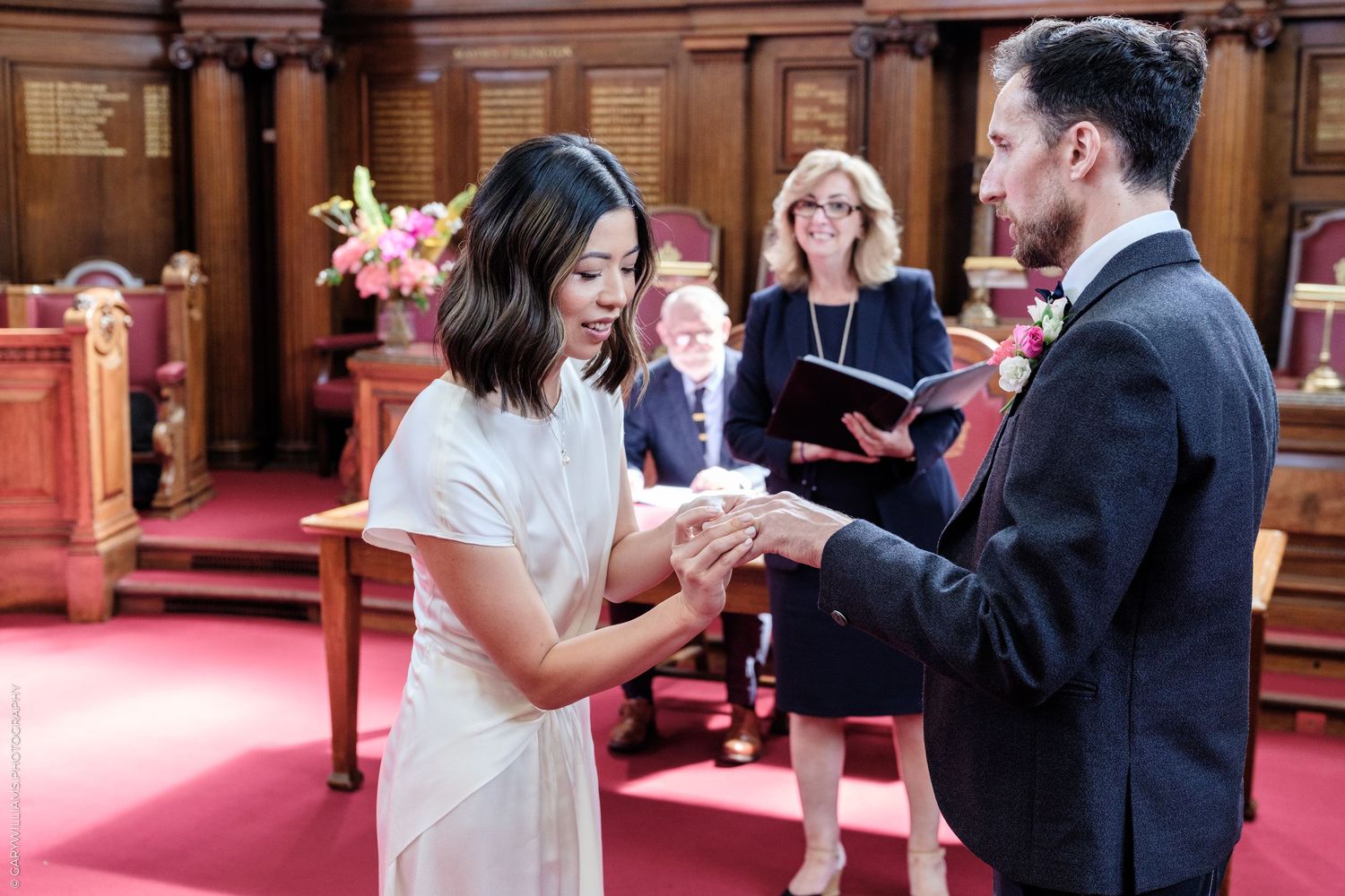 Couple exchanging rings during an intimate wedding ceremony in an elegant wood-paneled room with an officiant.