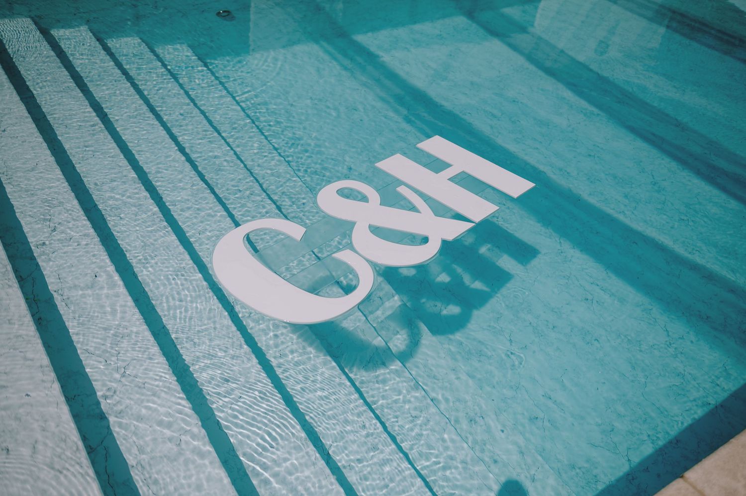 C&H logo letters submerged in a clear blue swimming pool, reflecting light on the tiled bottom.