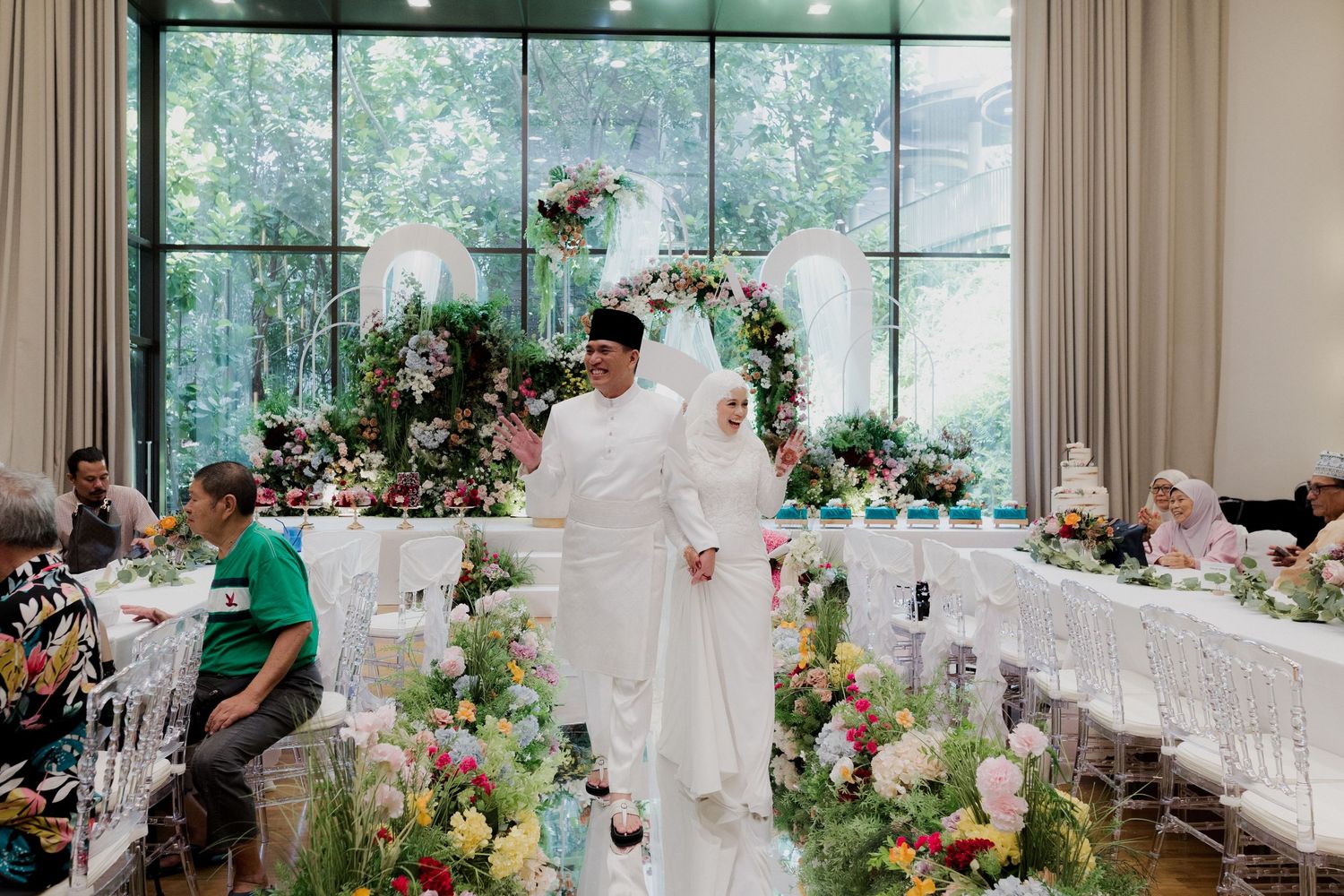 Muslim couple in white wedding attire walk through floral-decorated reception hall with guests seated at elegant tables.