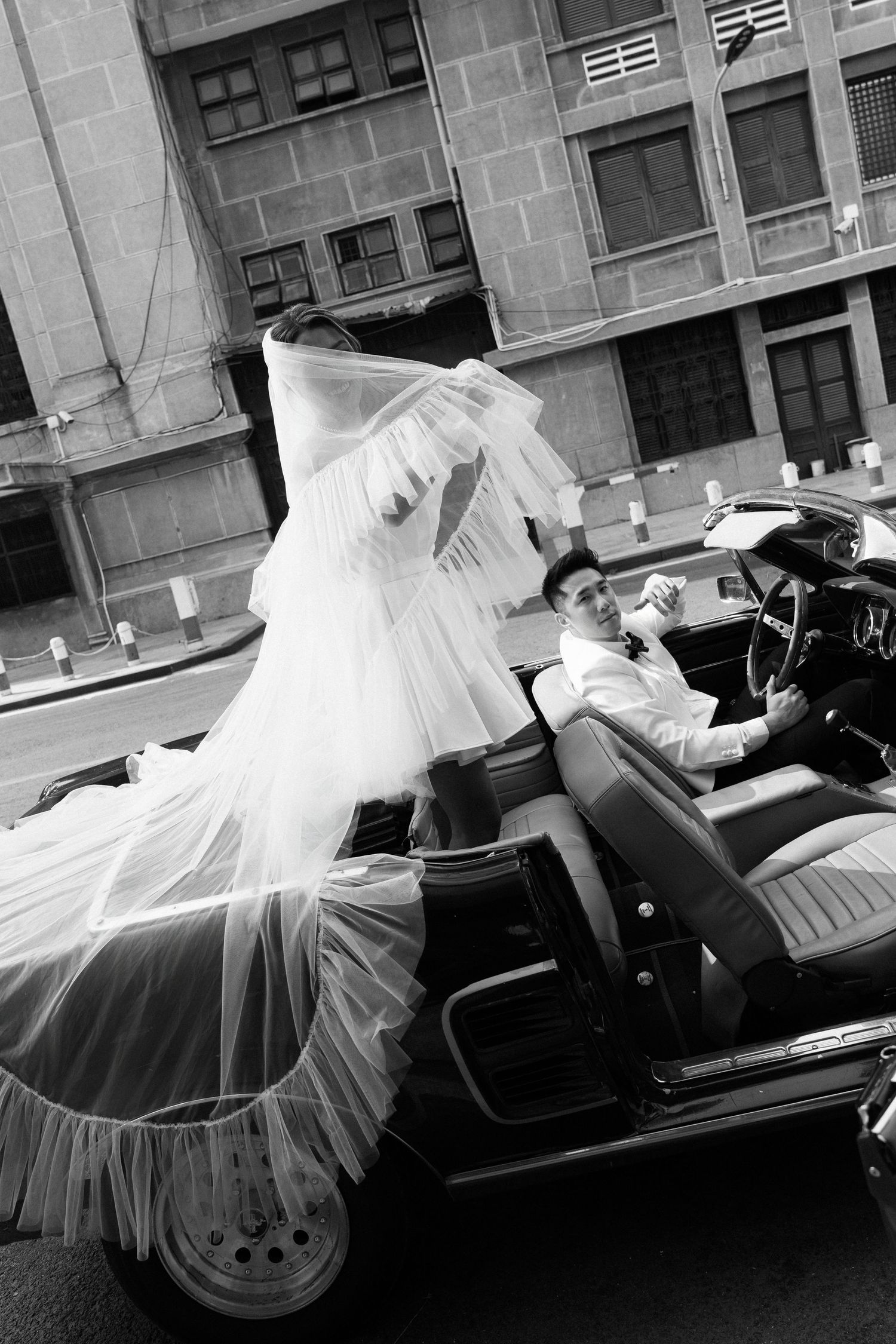 Black and white photo of a bride and groom in a classic convertible car on a city street, veil flowing in the wind.