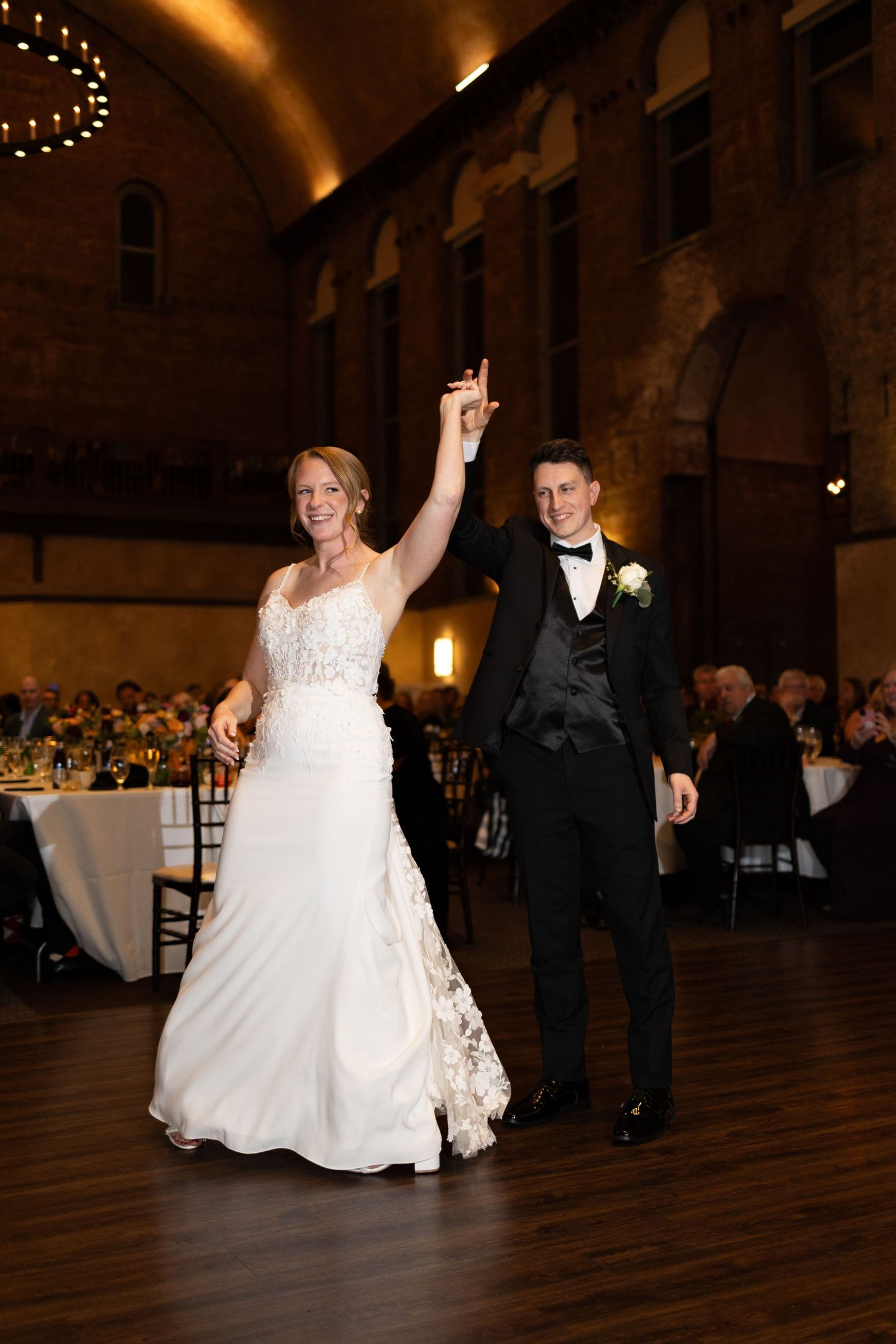 Bride and groom during first dance, bride raising hand joyfully in stunning white lace gown.