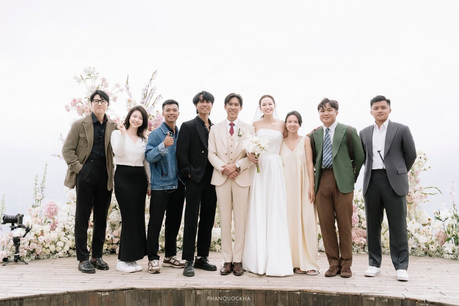 Wedding party group photo with bride in white gown and groom in beige suit, surrounded by guests outdoors.