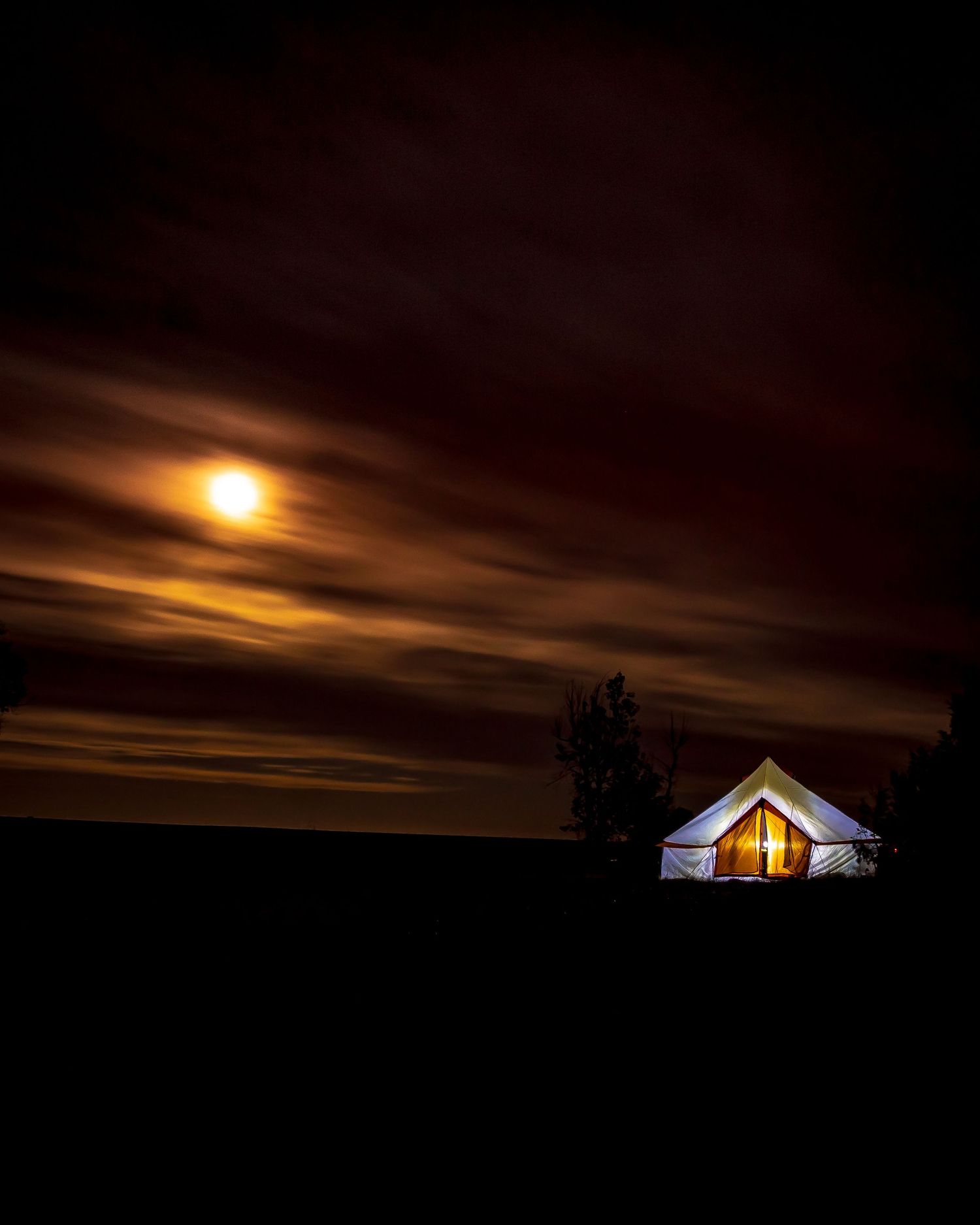 Glowing white bell tent illuminated at night under a dramatic moonlit sky with dark clouds and silhouetted trees.