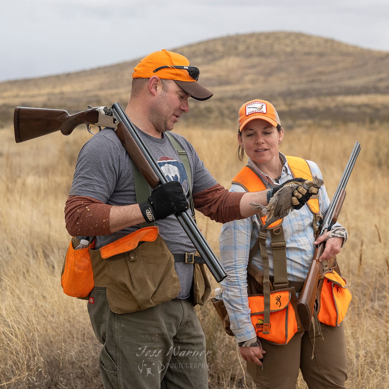 Two hunters in orange safety vests discuss firearms in a grassy field during a hunting trip.