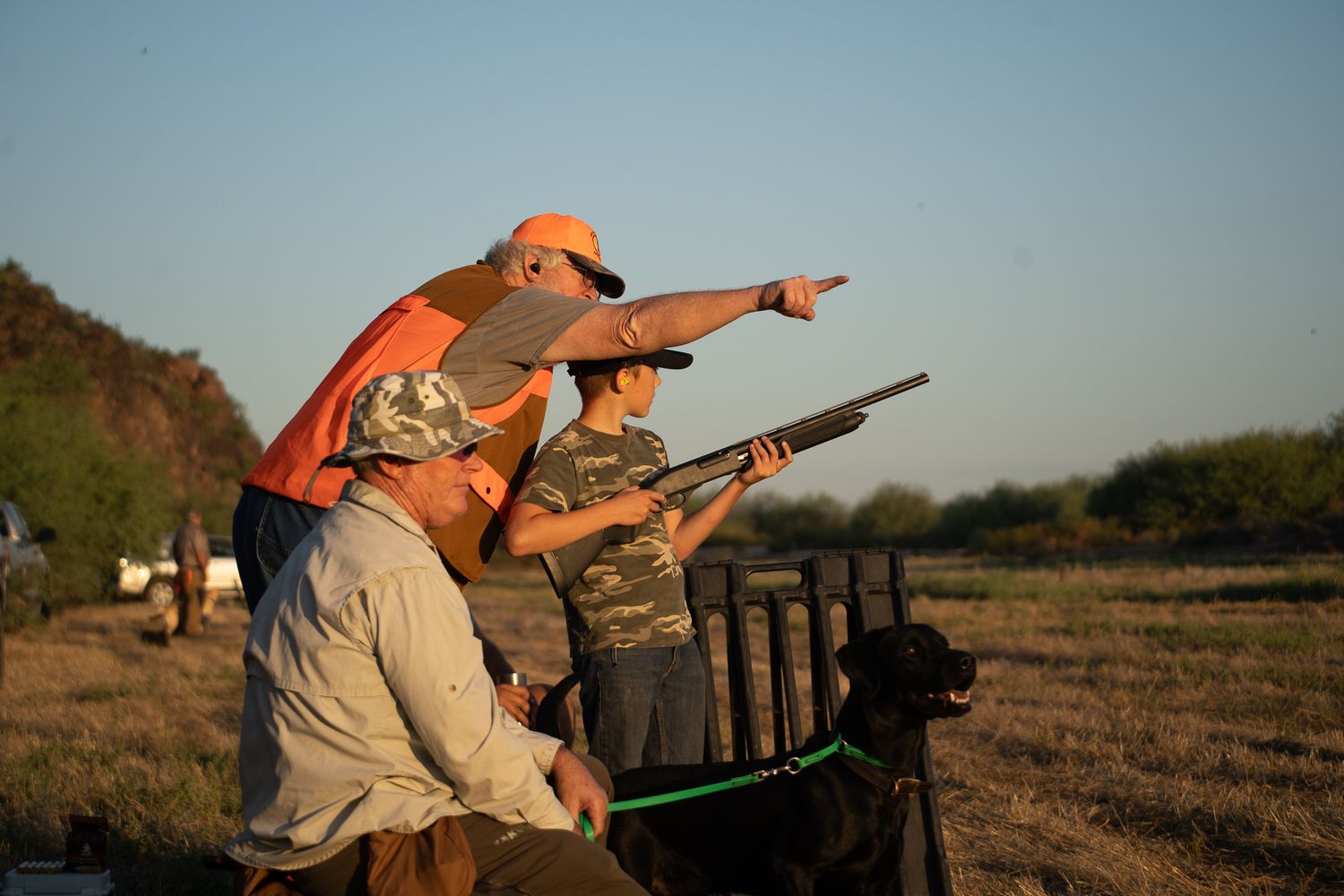 Three hunters with shotguns scan the field at golden hour, with a dog nearby during a bird hunting expedition.