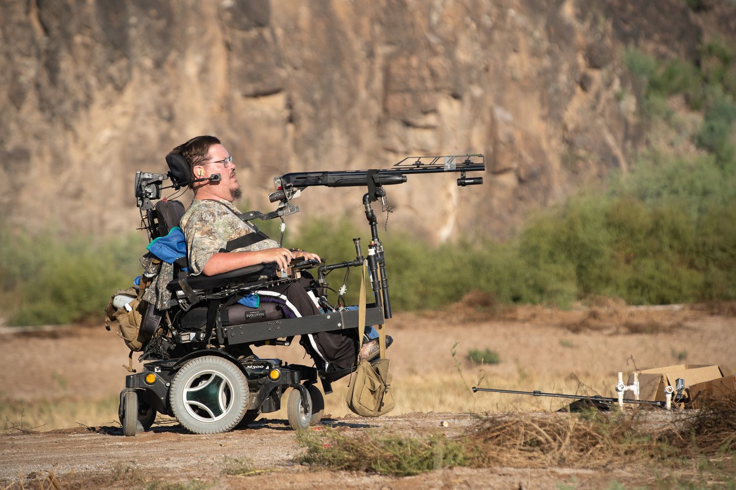 Person in a motorized wheelchair using an adaptive rifle mount for shooting in an outdoor rocky terrain setting.