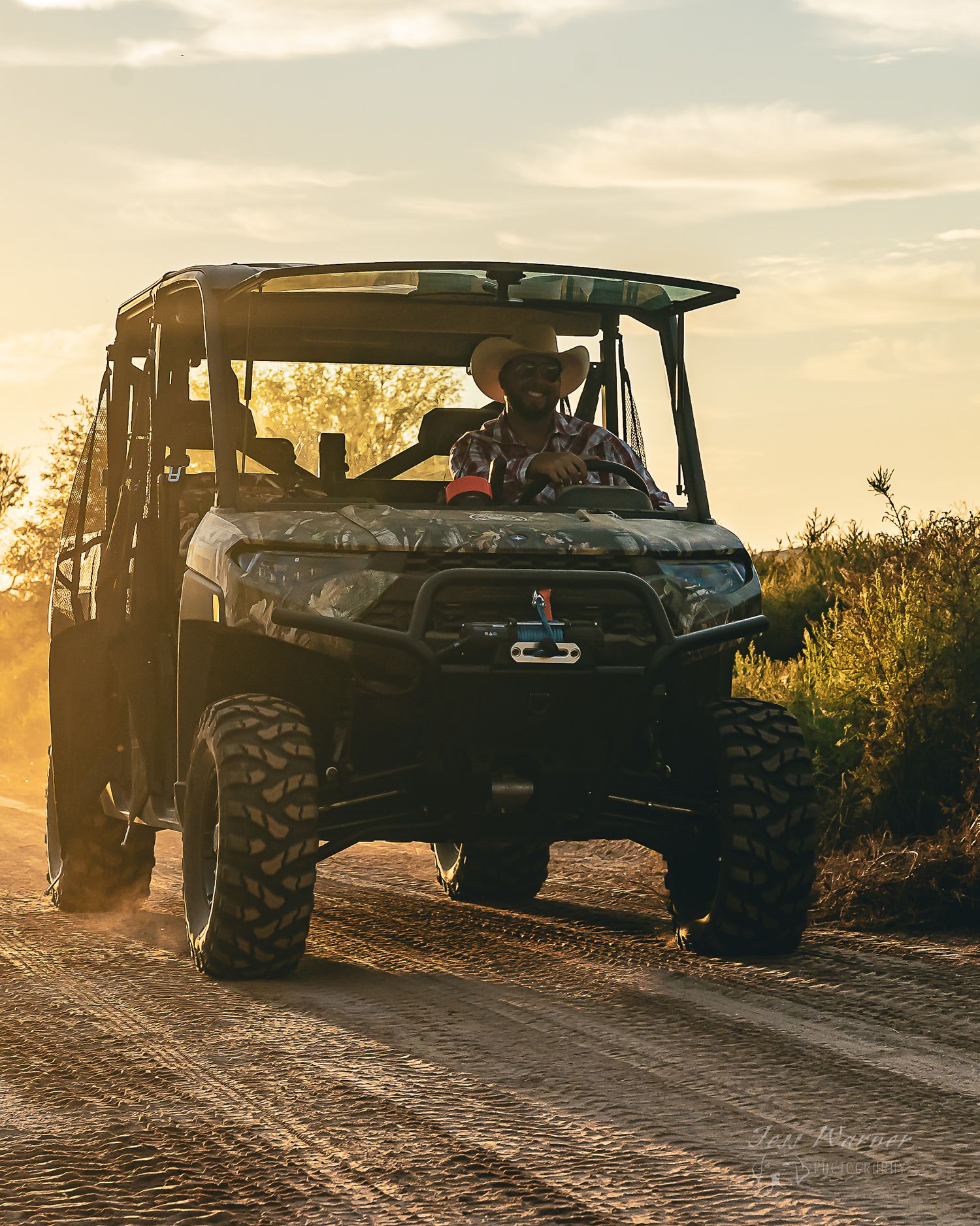 Person in cowboy hat driving a Can-Am Defender UTV on a dusty dirt road at golden sunset.