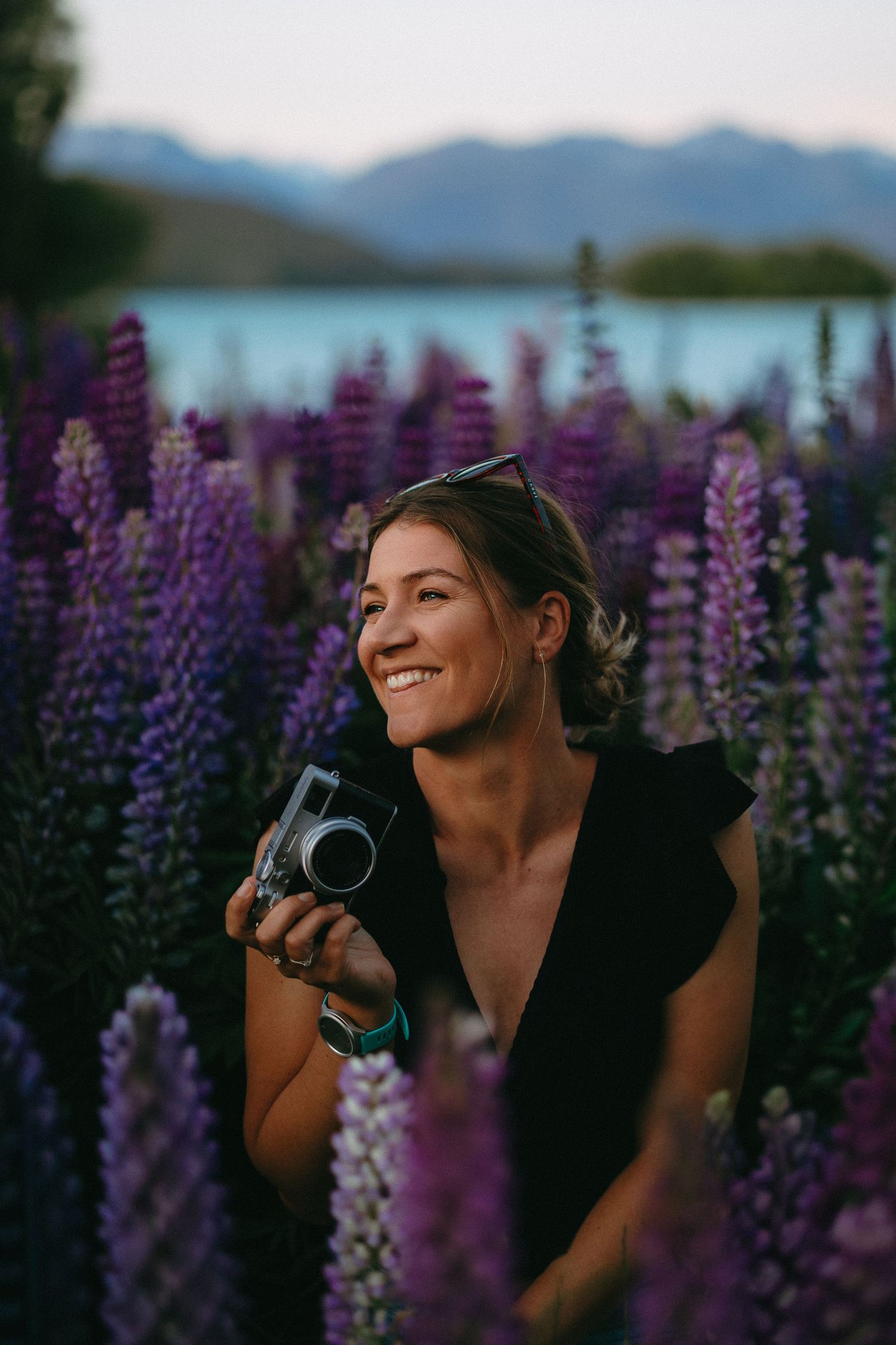 Smiling woman holding a vintage camera among purple lupine flowers with a turquoise lake and mountains in the background.