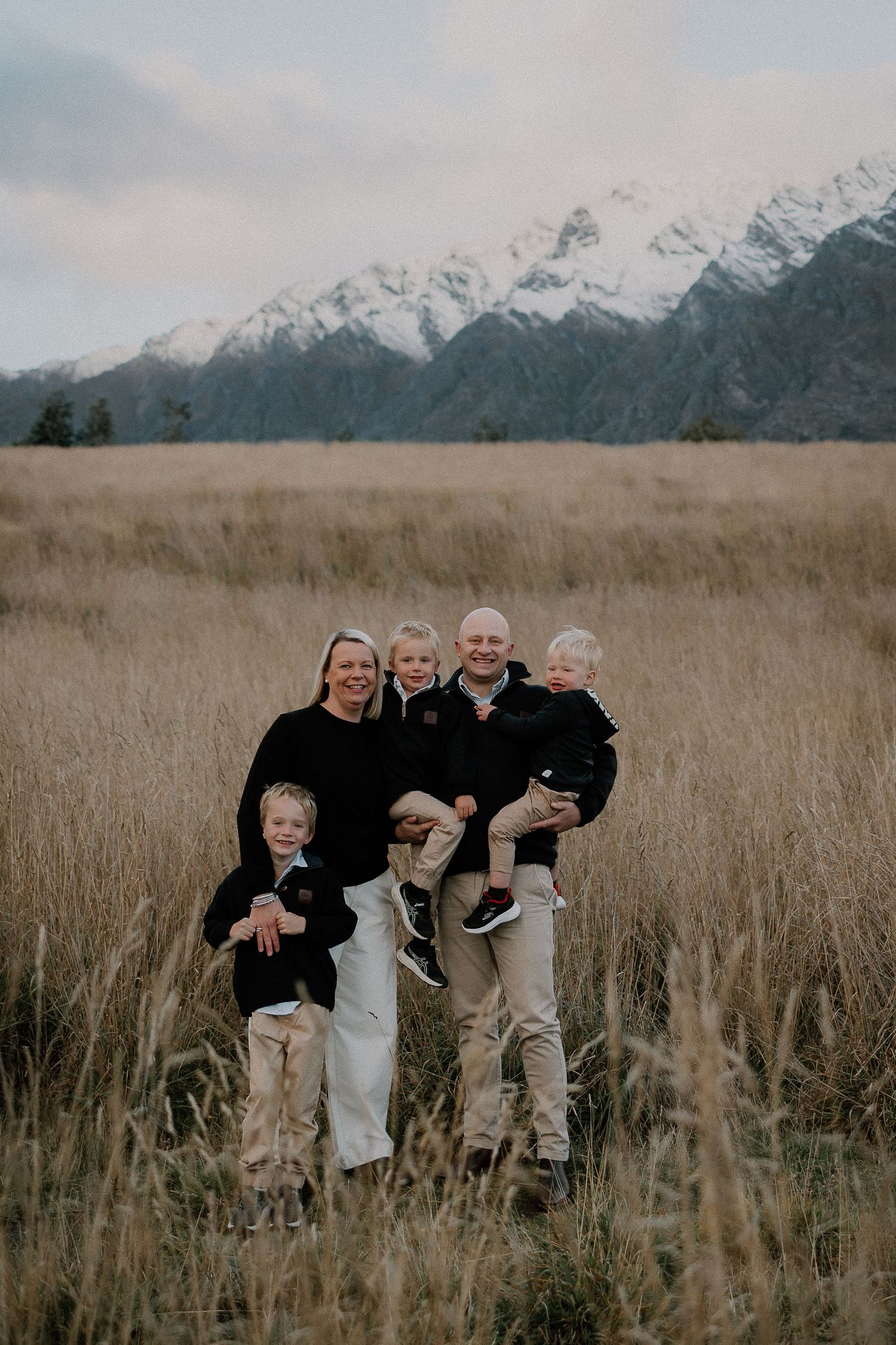 Family of five posing in golden field with snow-capped mountains backdrop, wearing matching black and beige outfits.