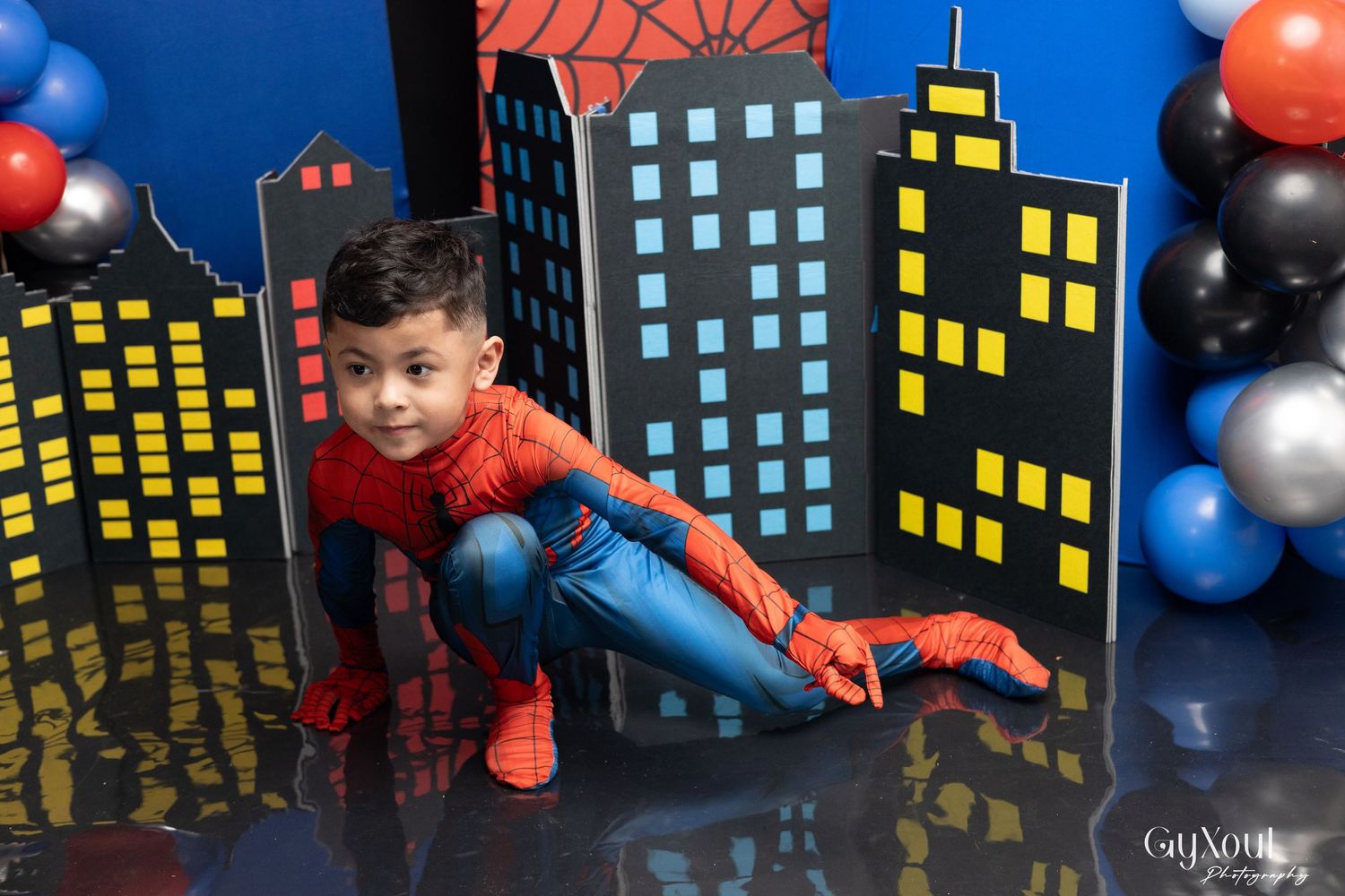 Boy in Spider-Man costume poses in front of cityscape backdrop with colorful balloons at birthday party.