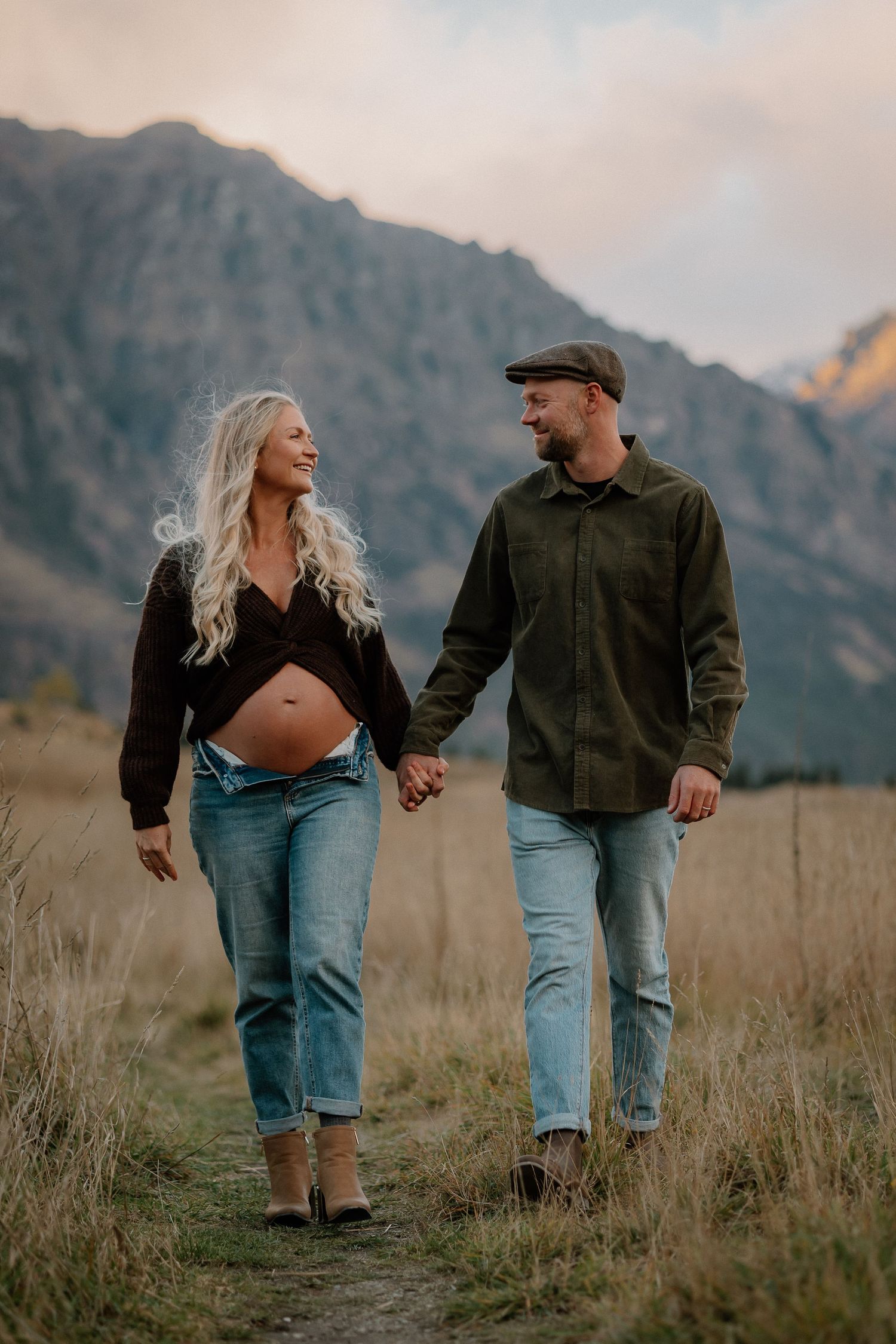 Pregnant couple holding hands walking on a mountain trail path, woman with long blonde hair and exposed baby bump.