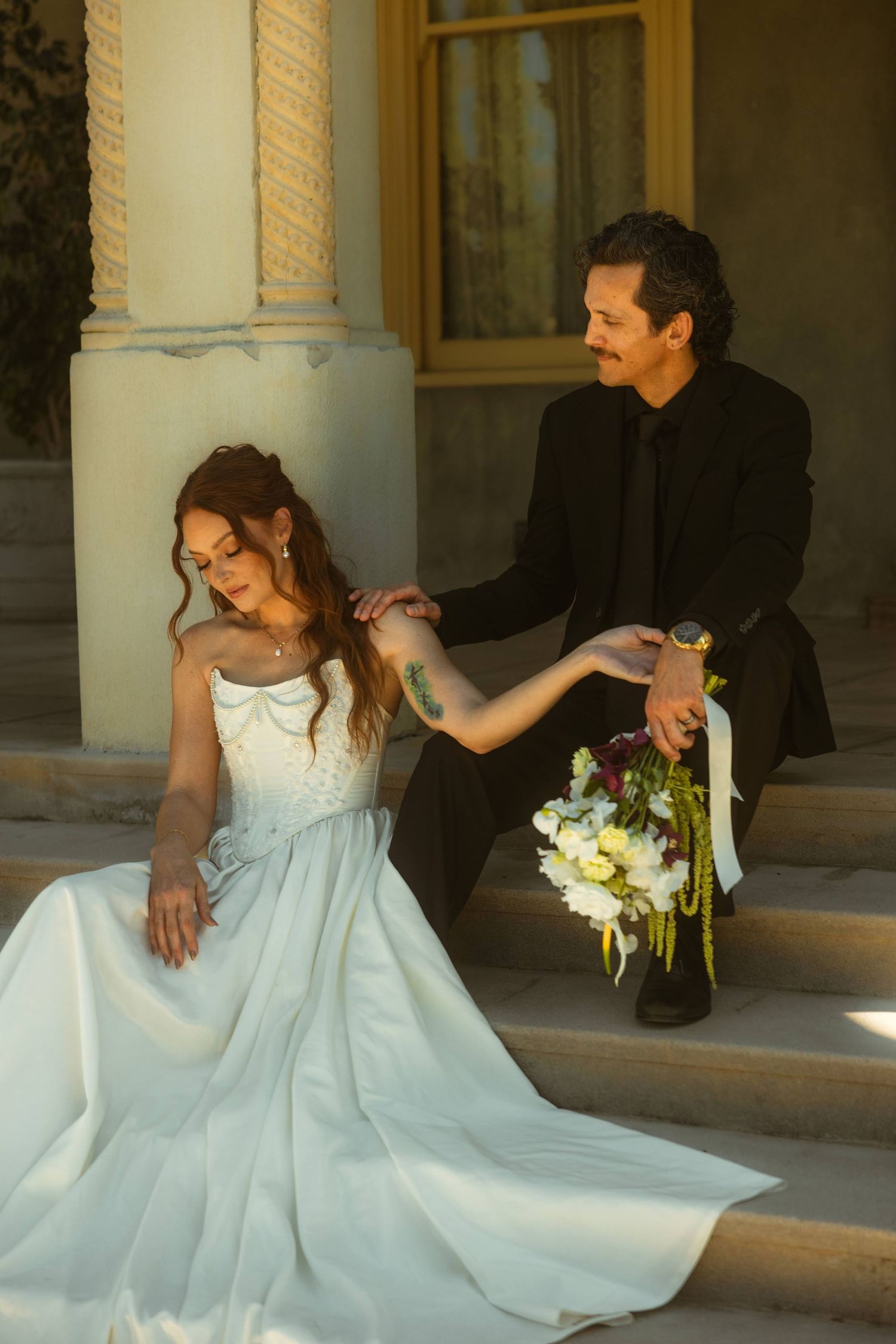 Bride in white gown sits on steps holding bouquet while groom in black suit holds her hand at the Kimberly Crest House wedding venue.