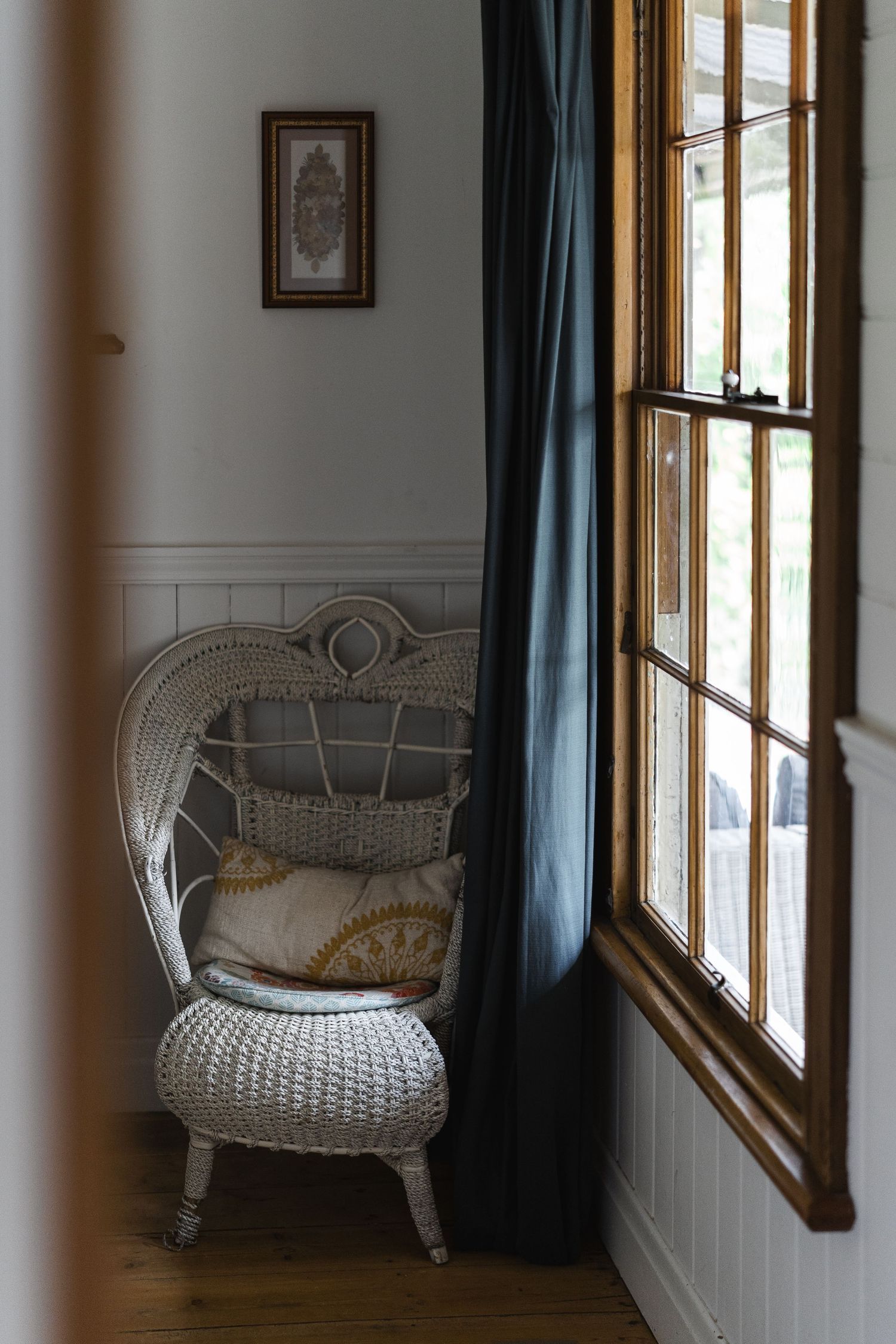White wicker peacock chair with decorative pillows beside dark curtains and a wooden window frame.

Rosey Knoll