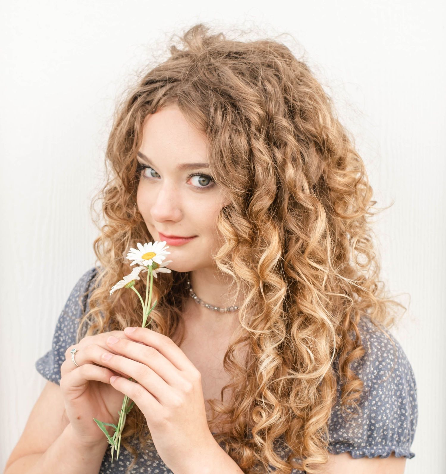 Young woman with long curly blonde hair holding a white daisy flower close to her face against a light background.