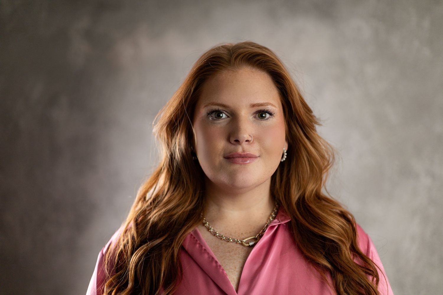Young woman with long red hair wearing a pink blouse and silver necklace poses against a gray backdrop.
