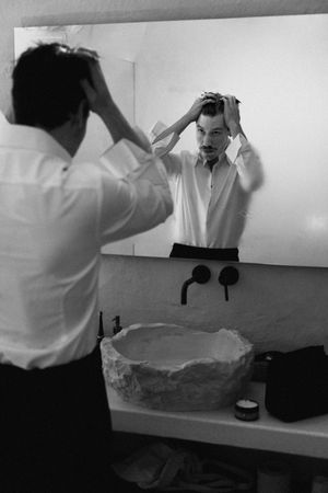 Man in white shirt styling his hair in a bathroom mirror, black and white photo with stone sink.