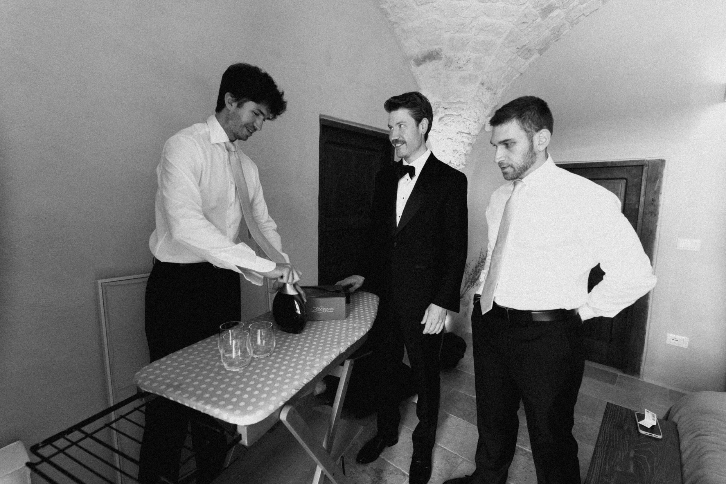 Three men in formal attire gather around an ironing board, one pouring champagne, in a black and white wedding preparation photo.