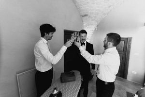 Three groomsmen in black and white photo raising glasses in a toast before a wedding ceremony indoors.