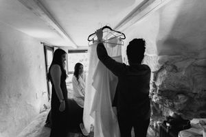 Black and white photo of bridesmaids helping hold up a wedding dress on hangers in a rustic stone room.