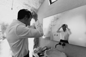 Black and white photo of a man fixing his hair in a bathroom mirror next to a sink.