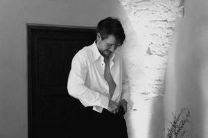 Black and white photo of a man in a white dress shirt buttoning up indoors near a textured white wall.