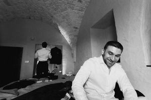 Black and white photo of two men in a vaulted stone room, one smiling in foreground, another standing by mirror.
