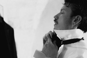 Black and white photo of a man in a white dress shirt adjusting his bow tie, preparing for a formal event.