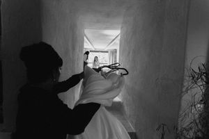 Black and white photo of a person holding a wedding dress in a narrow hallway with a bride visible in the background.