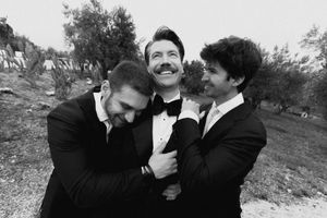 Three men in black tuxedos laugh and embrace outdoors in a black and white wedding photo.