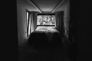 Black and white photo of a dimly lit bedroom with an unmade bed and a bright window in the background.