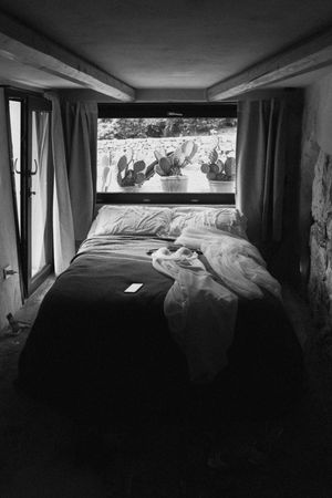 Black and white photo of an unmade bed with a large screen displaying cacti, flanked by curtains.