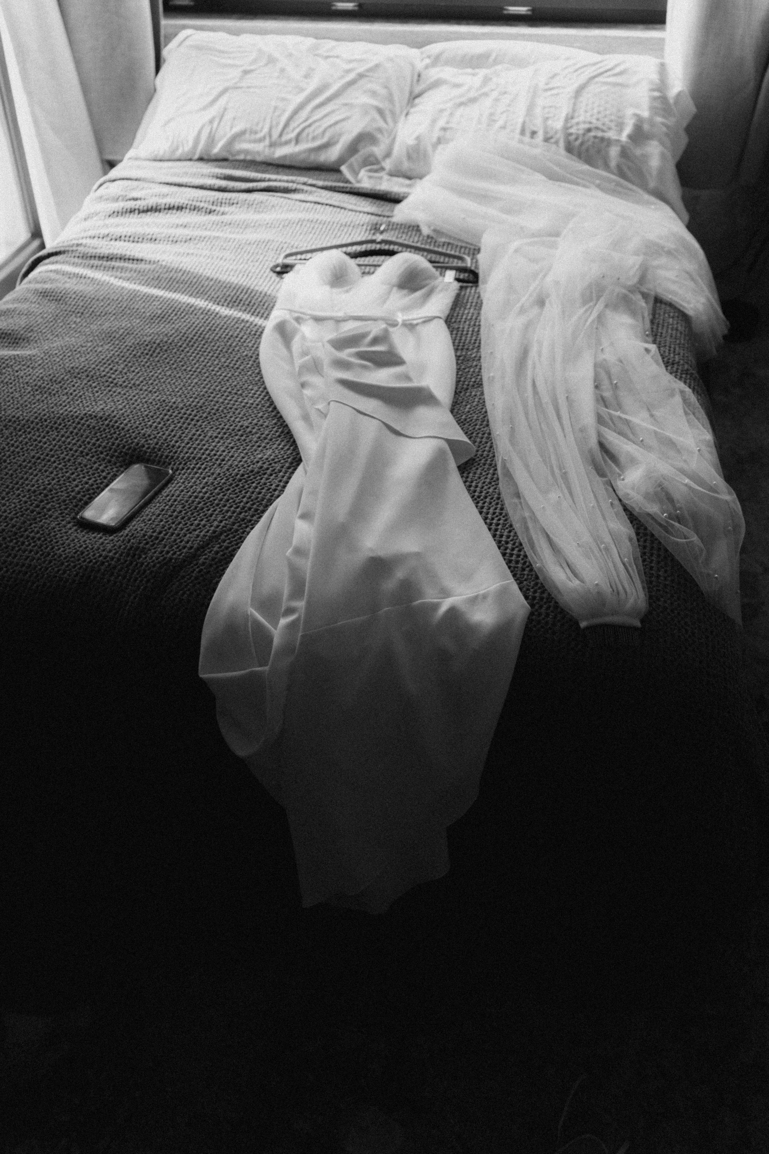 Black and white photo of a wedding dress laid on a bed with a veil beside it.