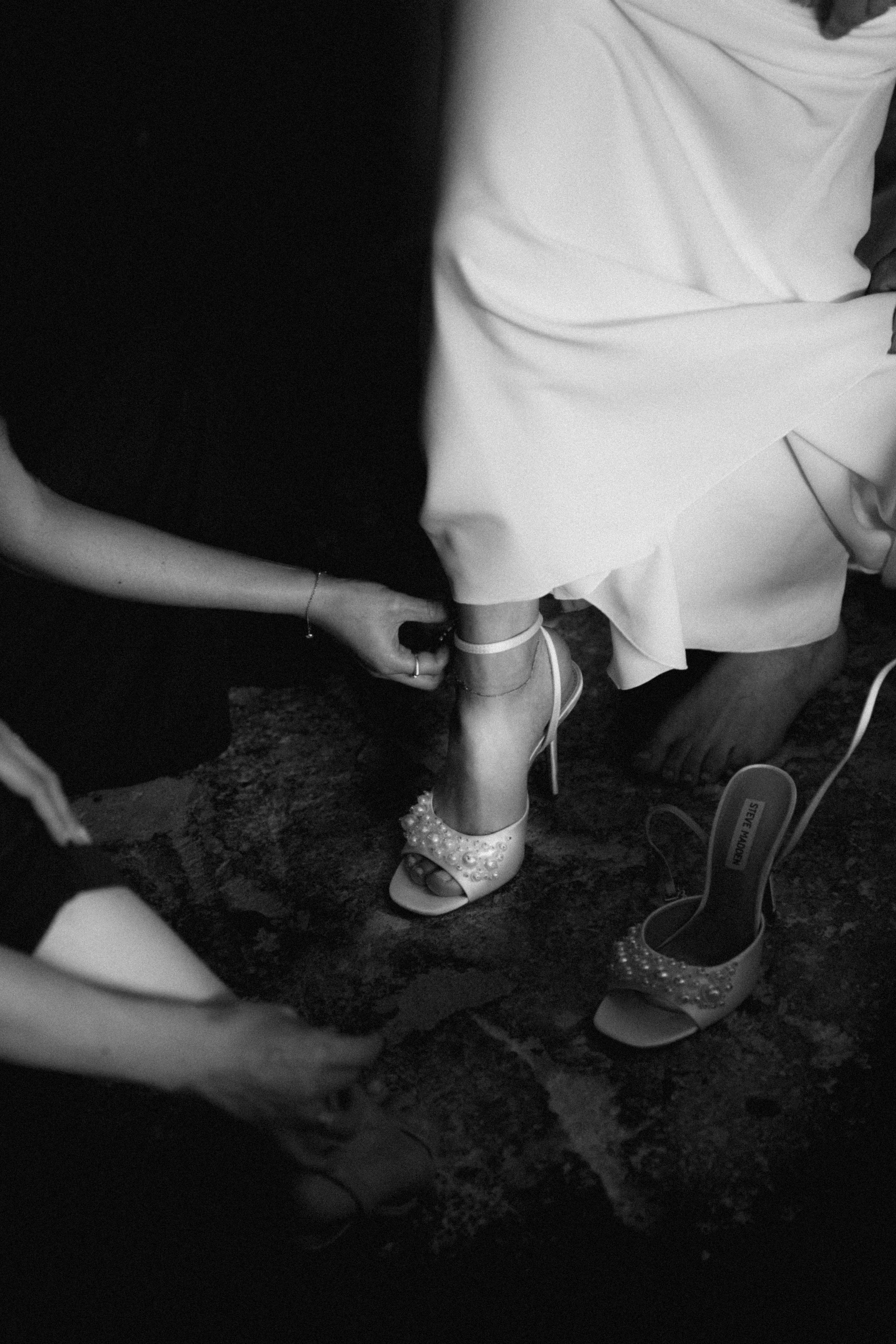 Black and white photo of a bride having her embellished heeled sandals fastened by a bridesmaid before the wedding.
