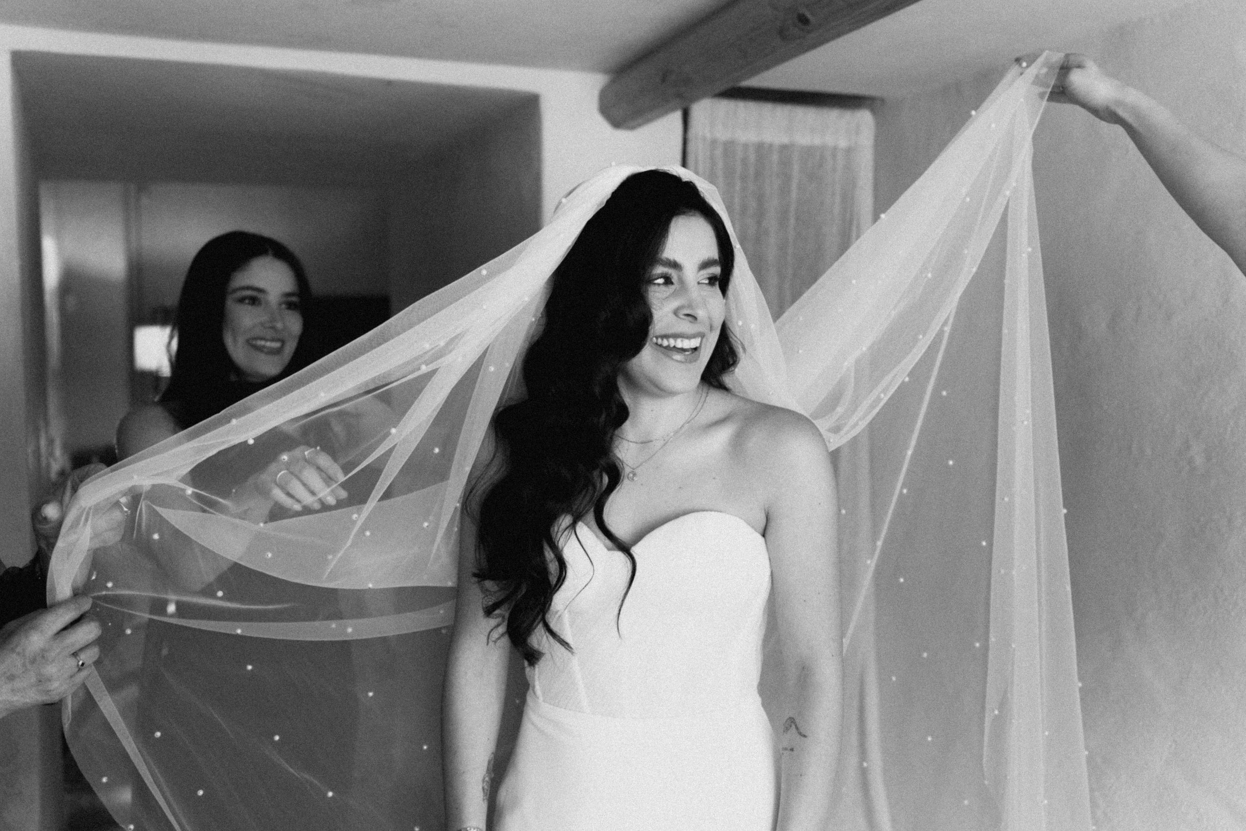 Black and white photo of a smiling bride in a strapless gown as her bridesmaid spreads her veil wide.