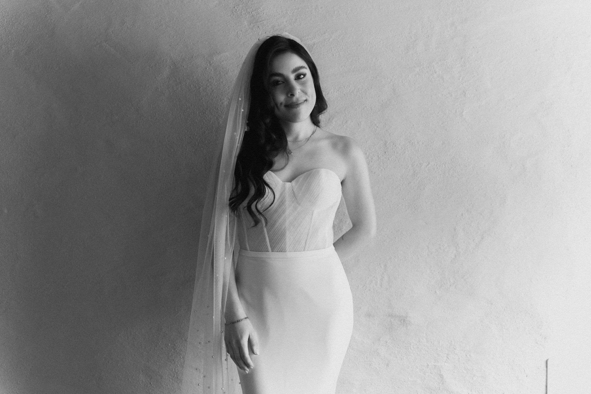 Black and white photo of a bride in a strapless gown and veil posing elegantly against a textured wall.