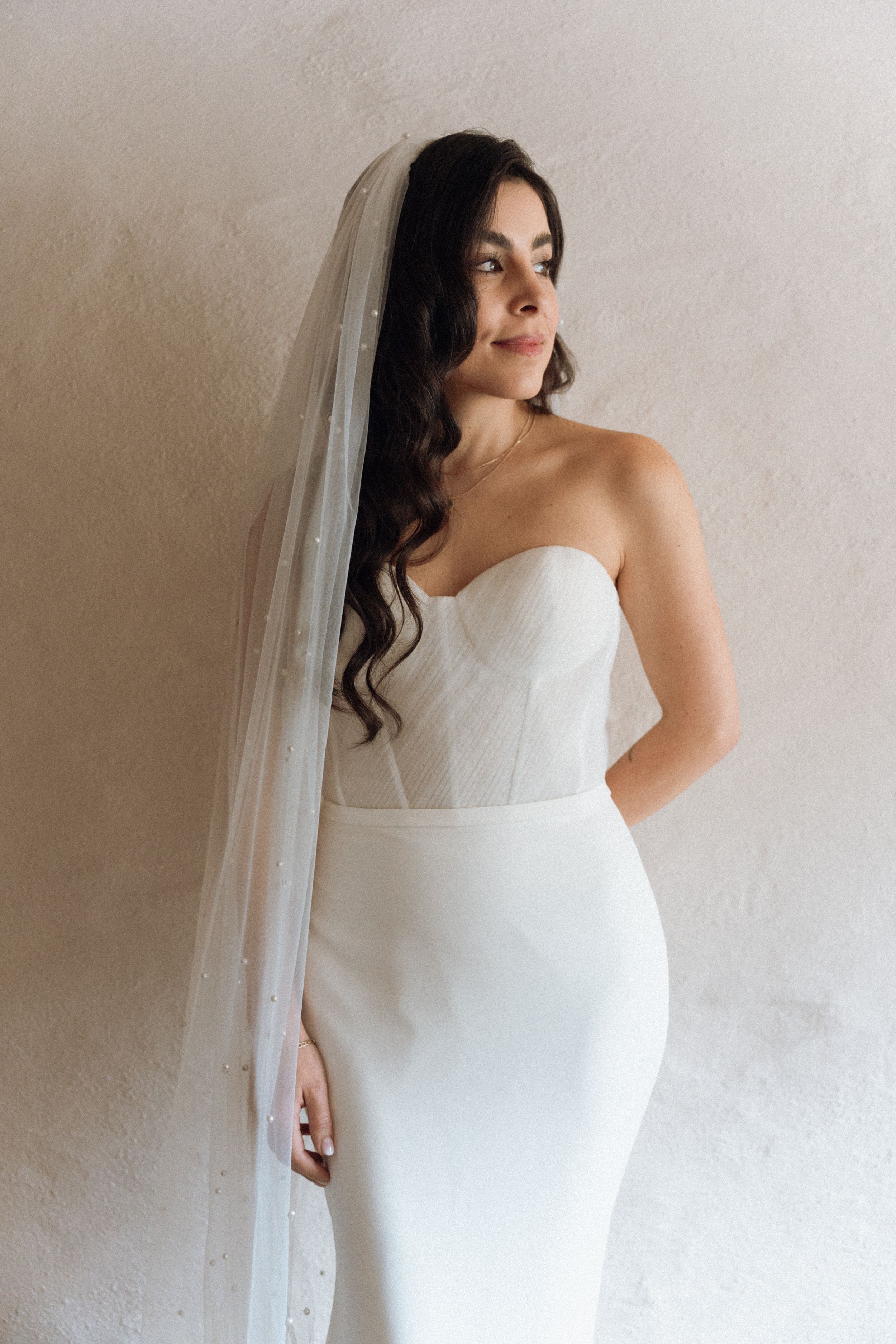 Bride wearing a strapless white corset gown with a pearl-embellished cathedral veil, posing against a neutral wall.