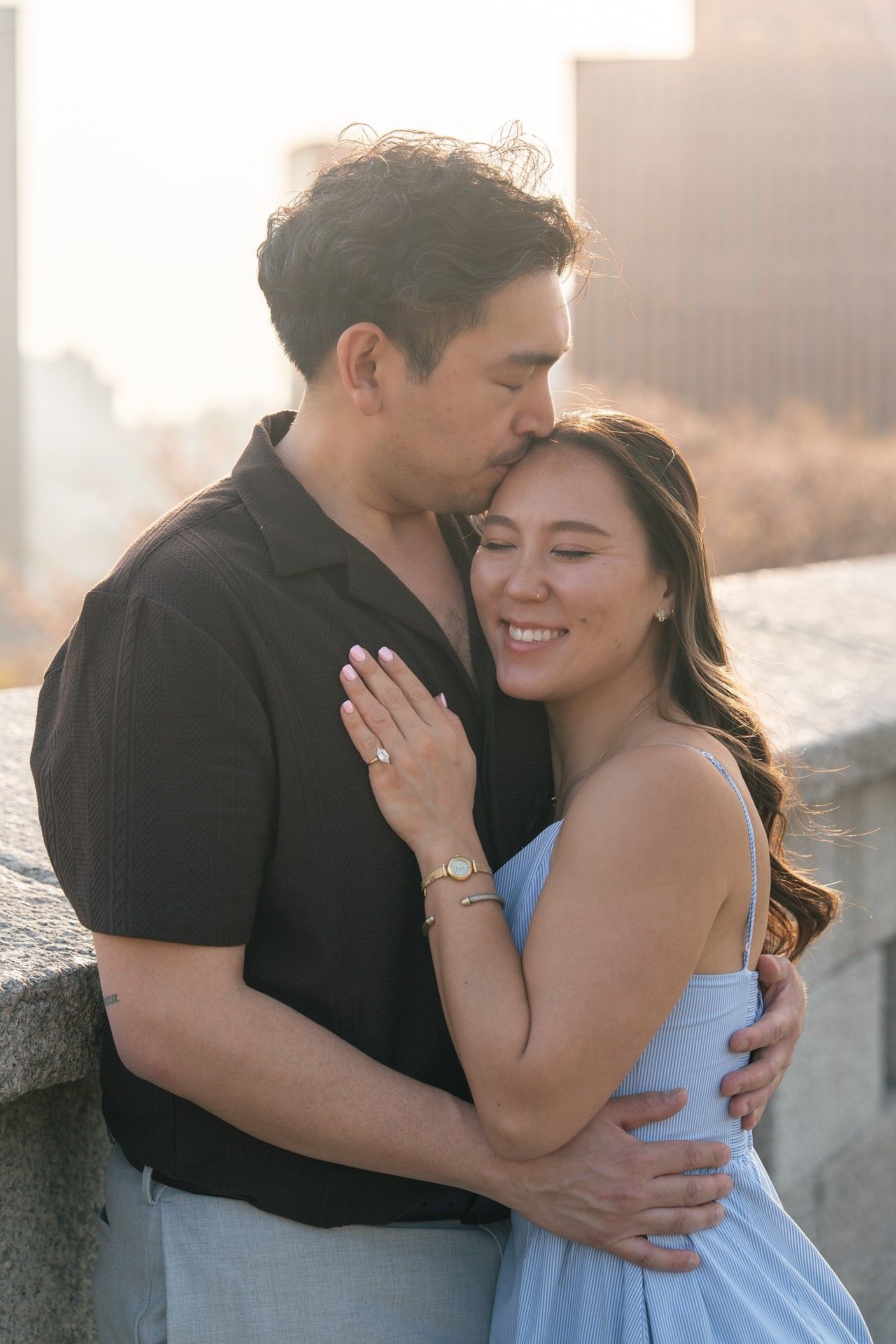 A couple shares a tender embrace at sunset, the man kissing the woman's temple as she smiles, wearing a blue dress.