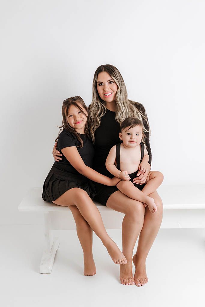 Mother in black dress poses with young daughter and baby boy on white bench in studio family portrait.