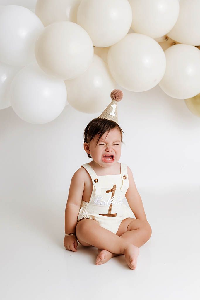 Crying baby in cream romper and birthday hat sits before white balloons at first birthday cake smash photo session.