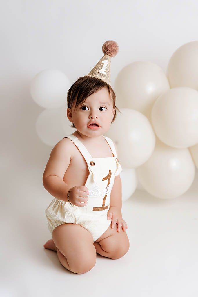 Baby's first birthday cake smash photo with cream romper, number one party hat, and white balloon backdrop.