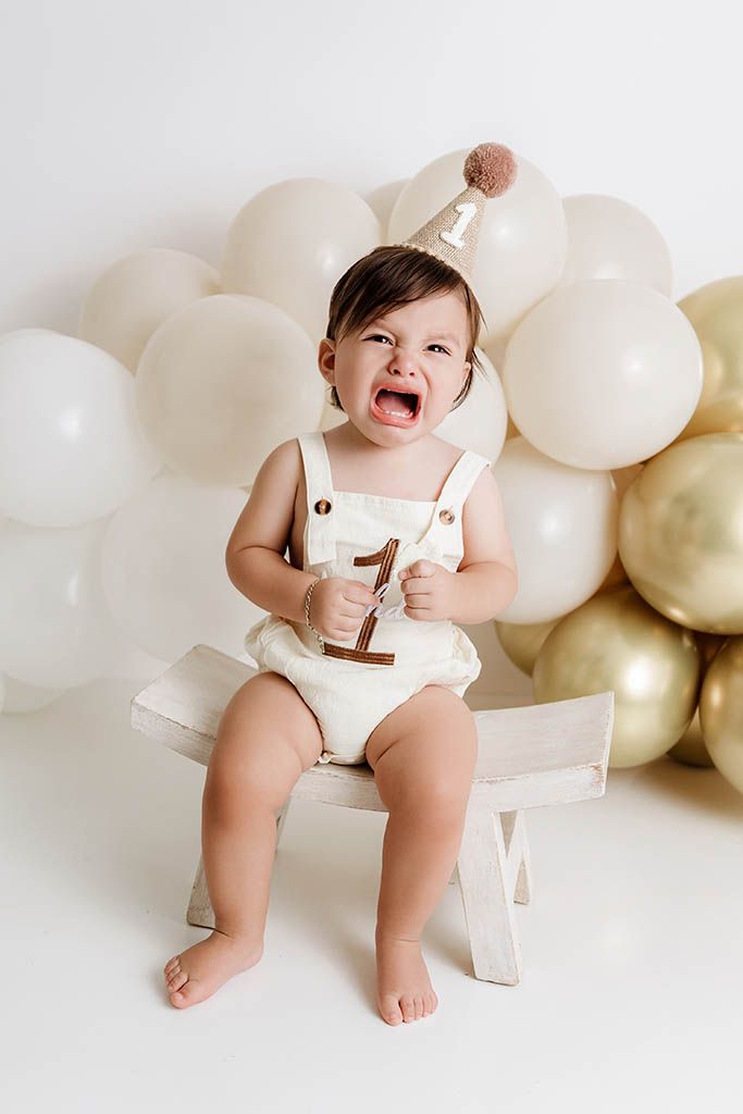 Crying baby in cream romper holding number 1, sitting on bench with gold and white balloons by Temescal Valley baby photographer.