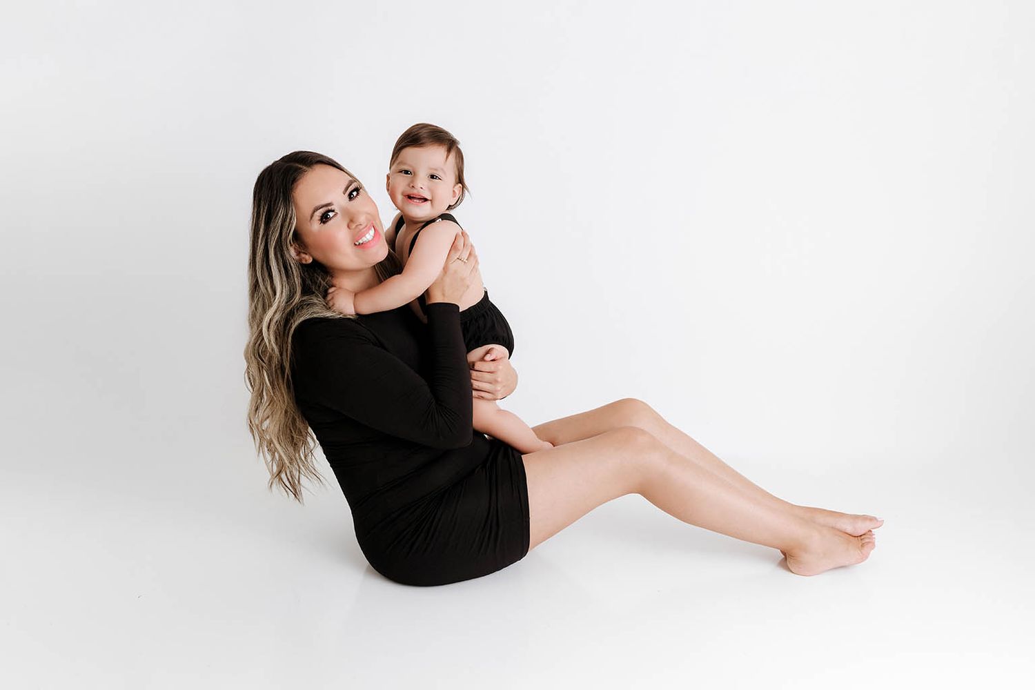 A mother in a black dress sits on a white studio floor, smiling while holding her happy baby girl.