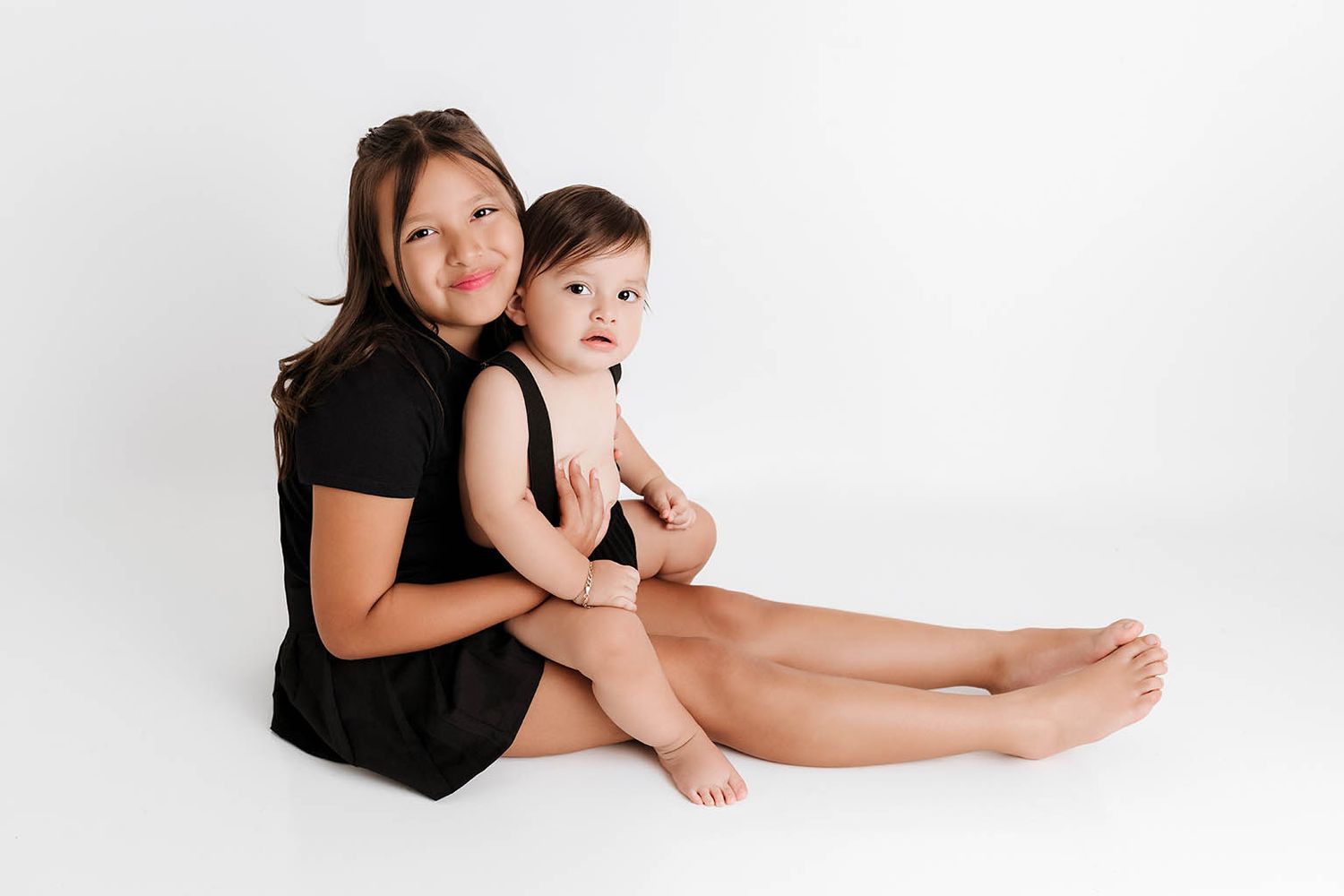Older sister in black dress hugging baby sibling during professional white studio photo session.